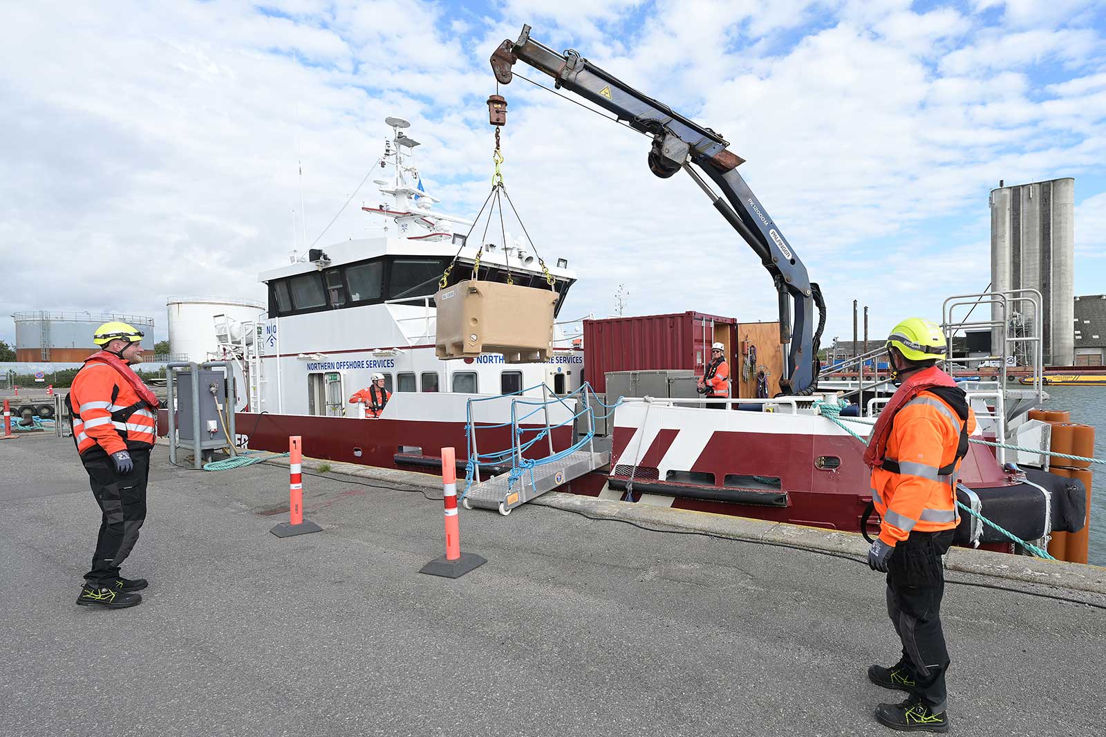 Two men in safety gear by a boat, with a crane lifting a box from the dock.