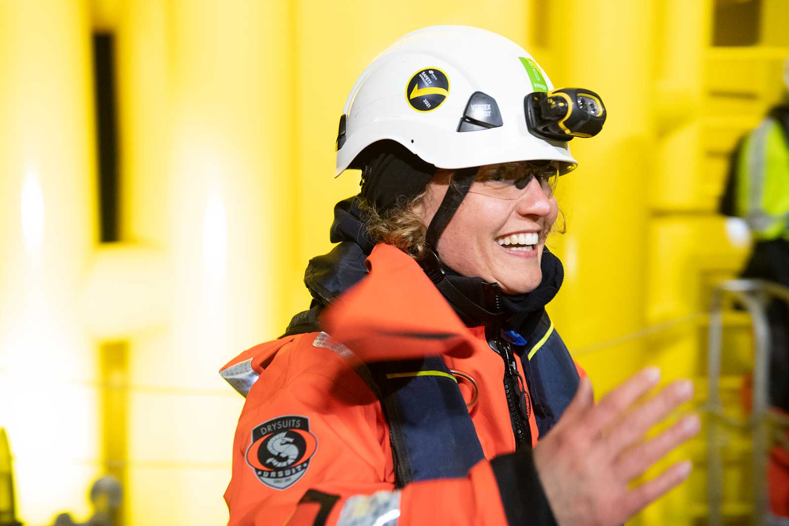 A person wearing an orange drysuit and helmet stands against a yellow background, gesturing with their hand.