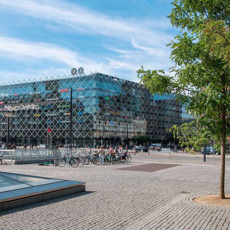 En moderne bygning med glasfacade og cykler parkert foran. En klar blå himmel og træer kan ses i forgrunden.