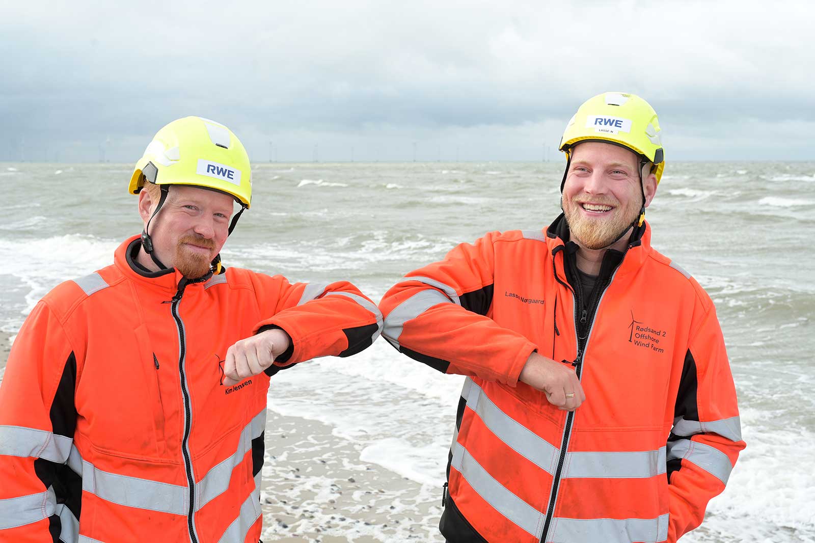 Two individuals in orange safety gear stand by the sea, displaying their arms with palms facing inwards.