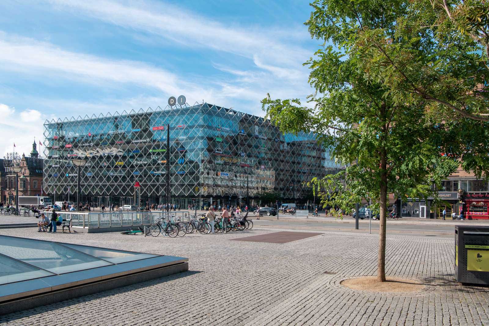 Moderne bygning med glasfacade og cykler i forgrunden. Blå himmel og træer ses i området.
