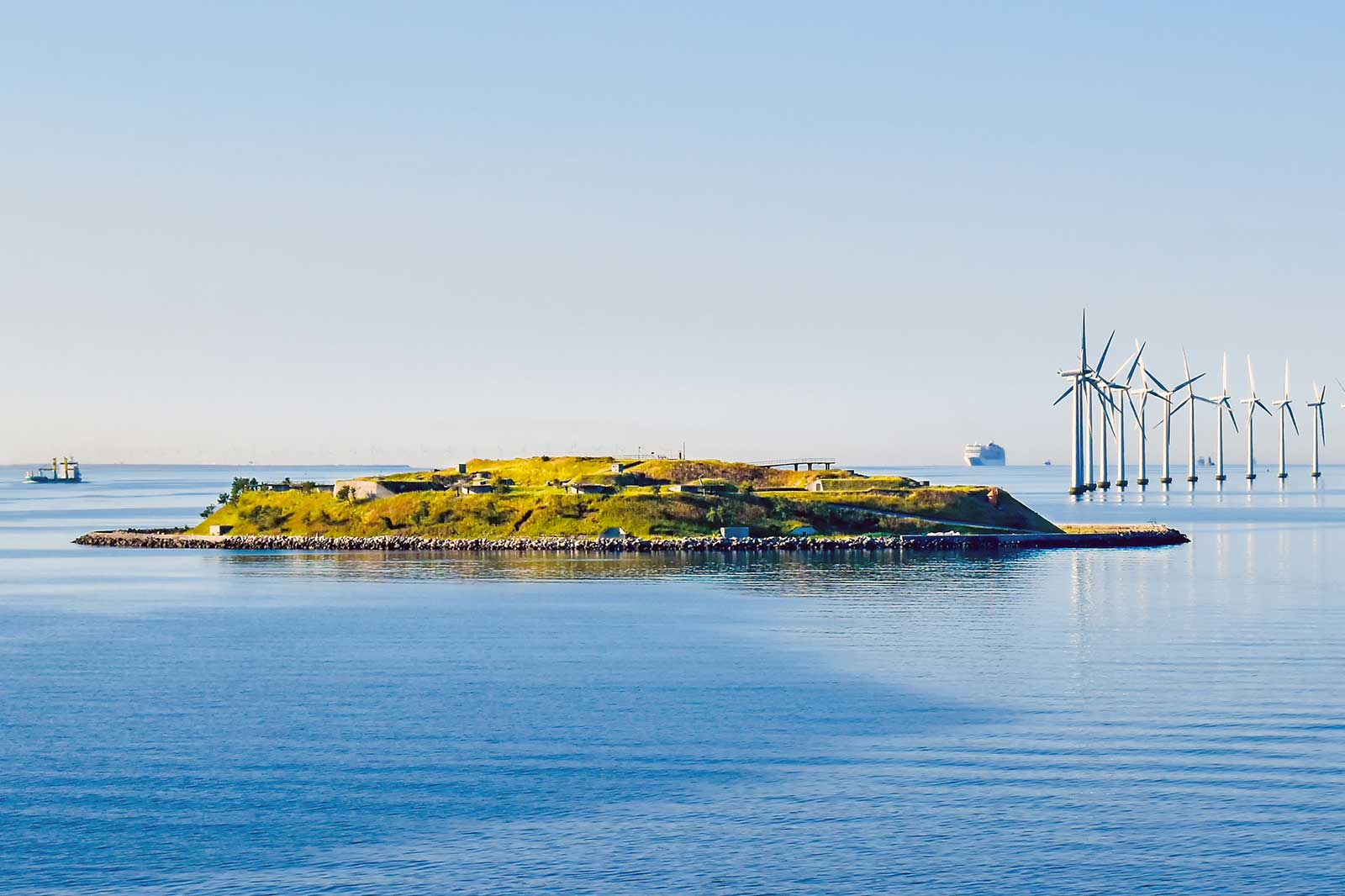 A small island with grass and buildings, surrounded by calm water and wind turbines in the background.