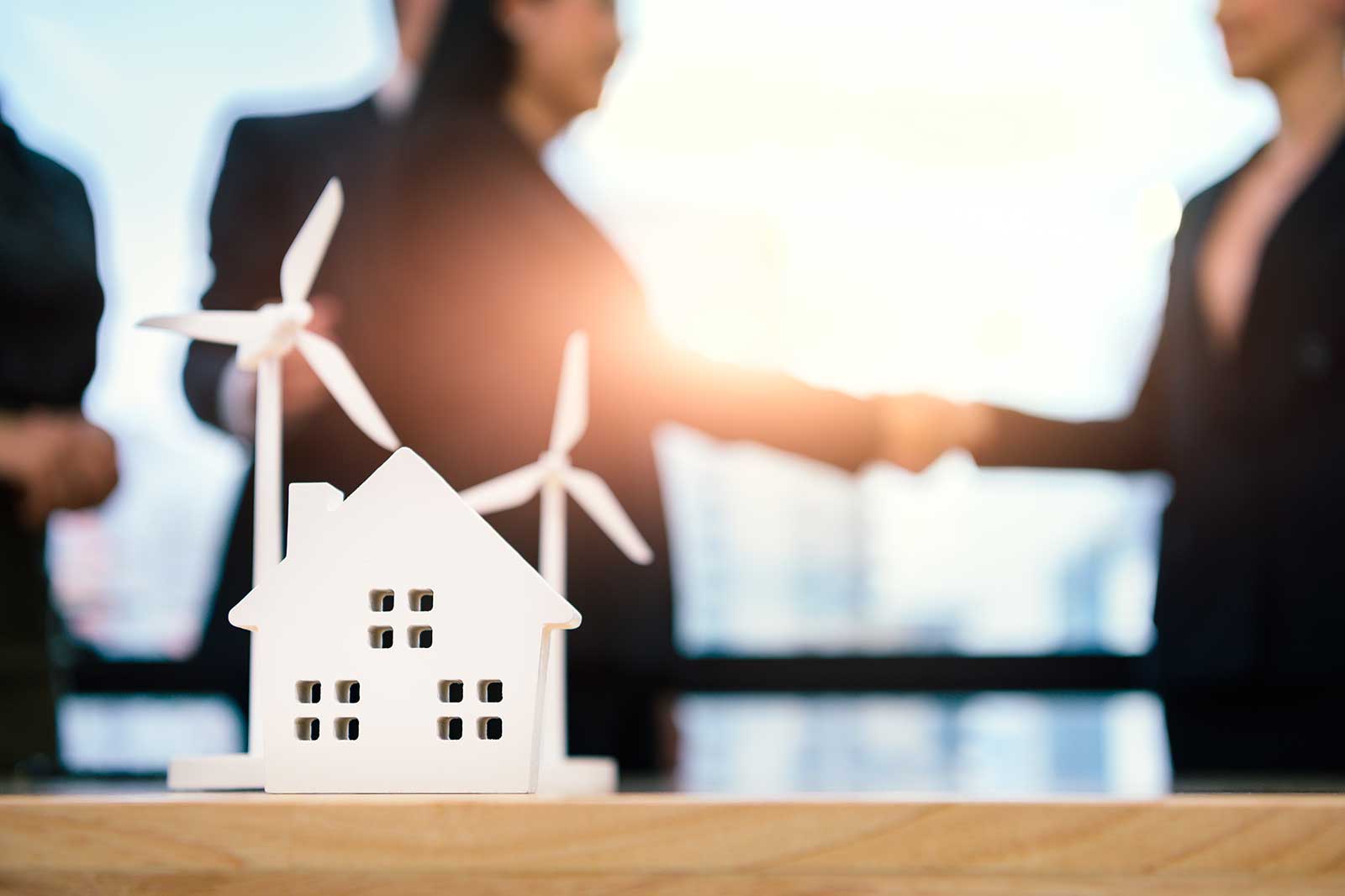 A model house with wind turbines in the background. Two people are shaking hands in a bright office environment.