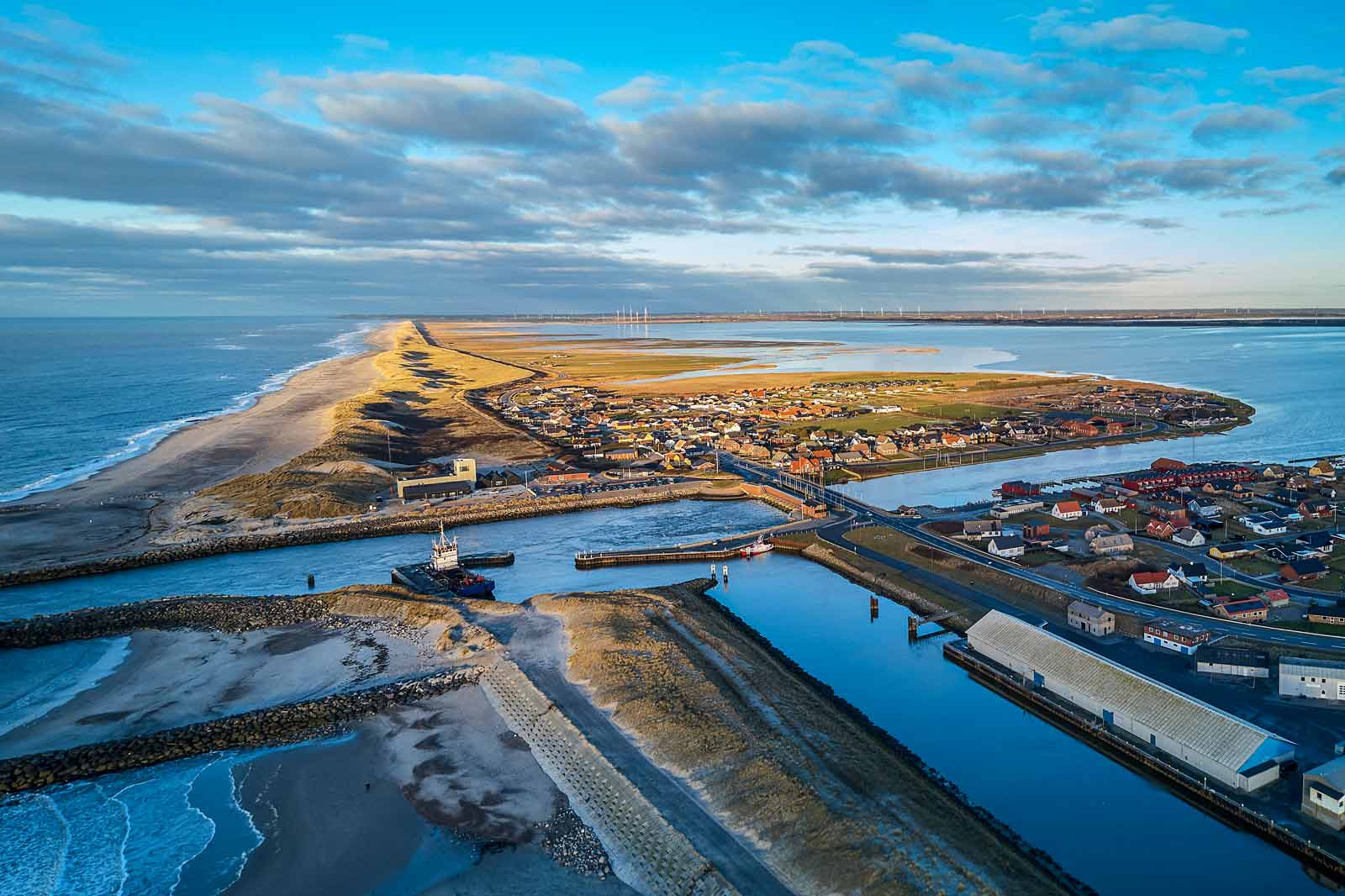 Luftfoto af et kystlandskab med en strand, en landsby og vandveje under en blå himmel med spredte skyer.