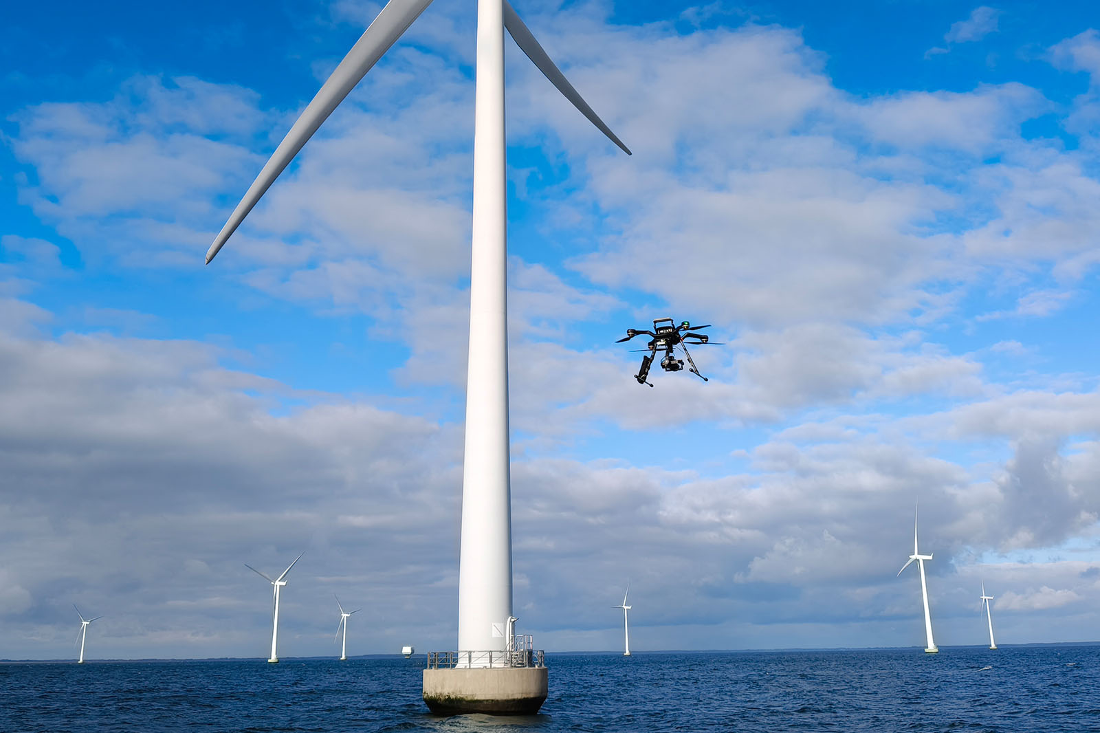 En drone flyver ved siden af en vindmølle over havet, med flere vindmøller i baggrunden under en blå himmel.