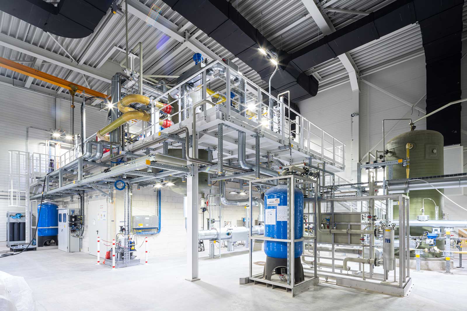 A large stainless steel processing structure with platforms and intertwined pipes dominates a bright white industrial hall, with blue tanks and yellow insulated piping visible.