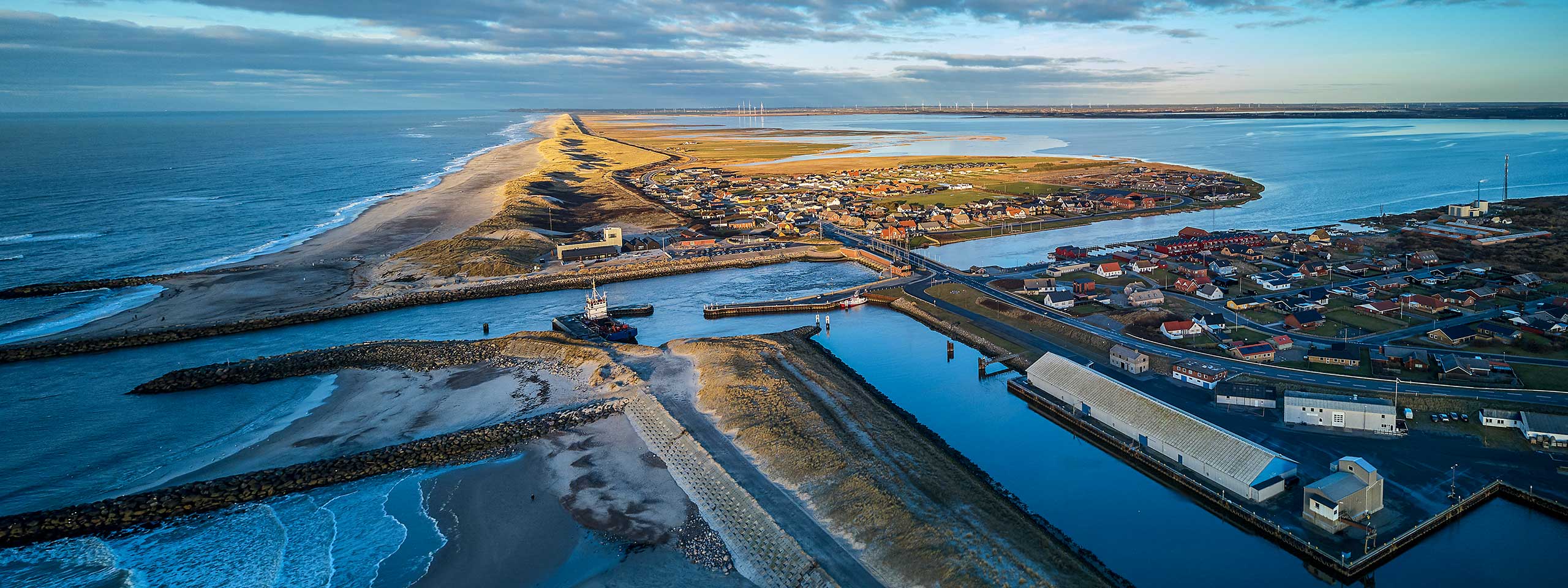 Luftbillede af en kystlinje med huse, en havn og bølger ved havet.