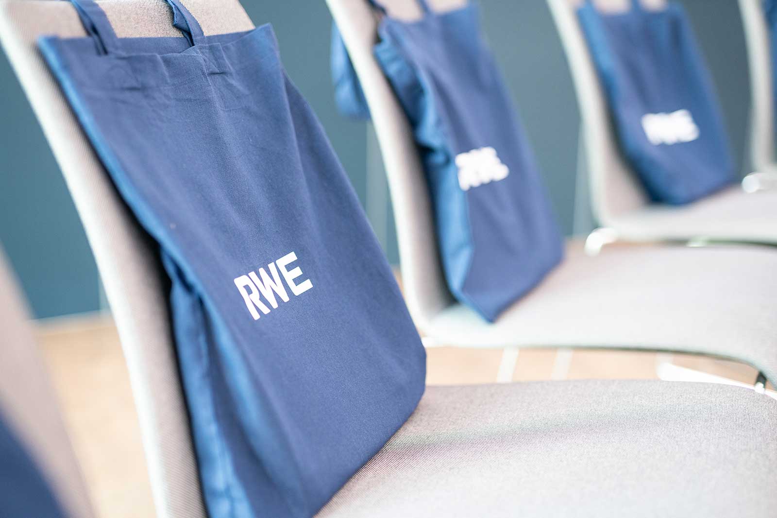 A navy blue fabric tote bag with the white text "RWE" hangs over a gray upholstered conference chair, repeated along a row.