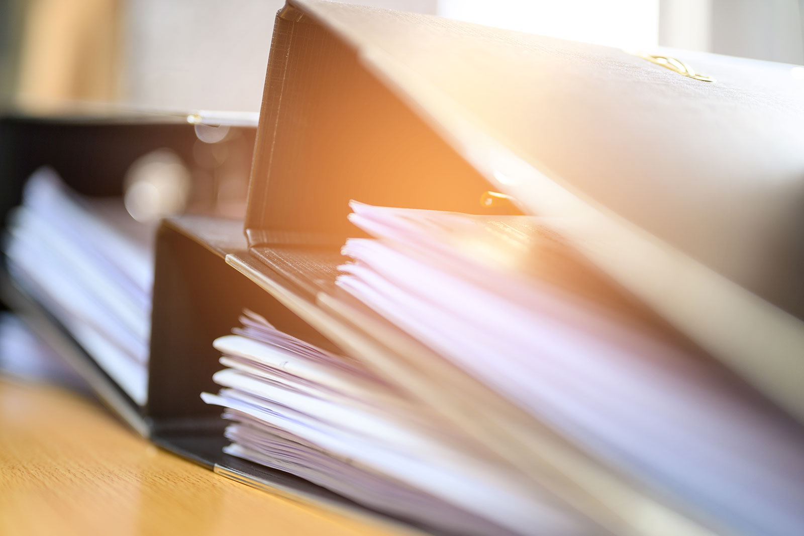 A black binder lies open on a wooden desk with white papers fanned under warm sunlight, giving a calm, orderly impression.