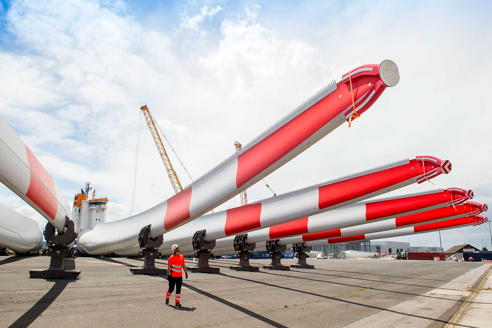 A person in work attire walks among large wind turbine blades with red and white stripes, parked at a harbour.