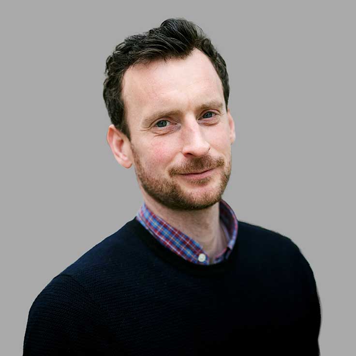 A person with dark hair, wearing a navy sweater and a checkered shirt, stands against a grey background.