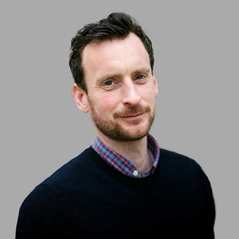 A person with dark hair, wearing a navy sweater and a checkered shirt, stands against a grey background.