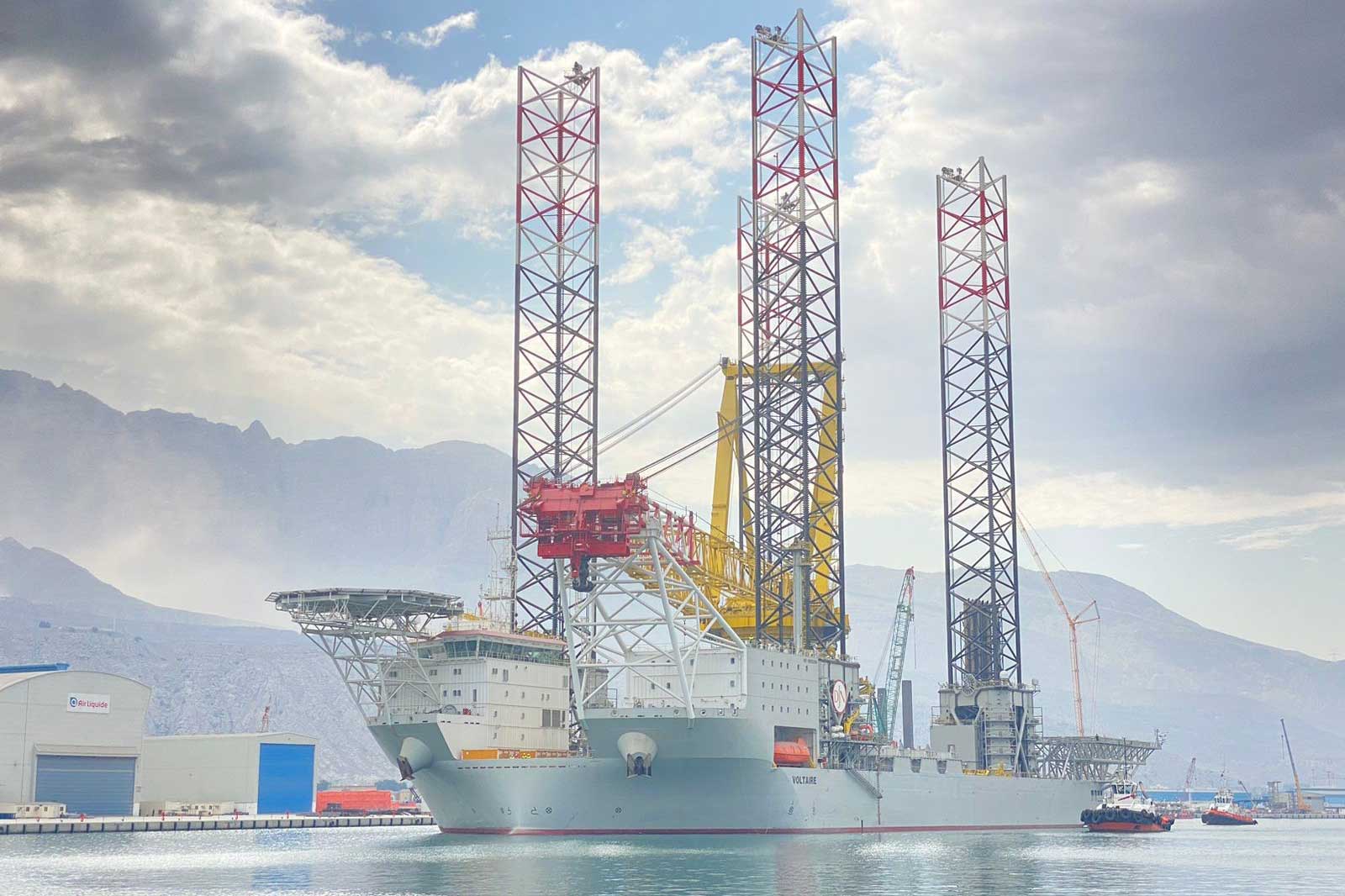 A drilling ship with tall legs docked in the harbour, surrounded by mountains and other vessels.