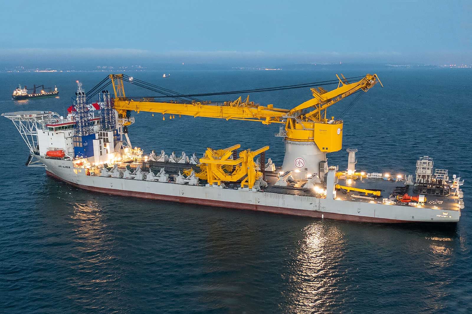 A large offshore supply vessel with a yellow crane, illuminated at night, floating in the sea with distant ships in the background.