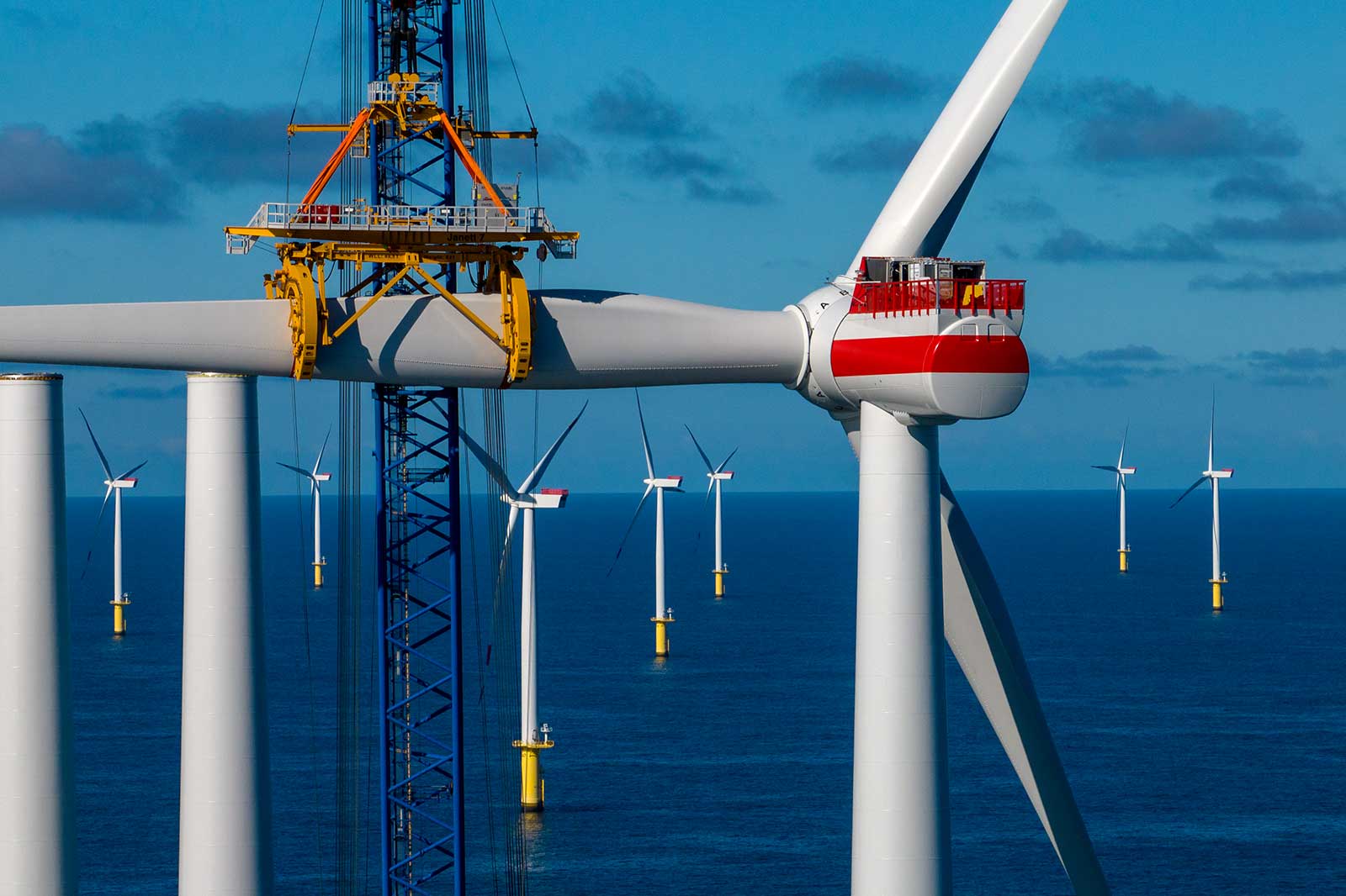 A close-up of a wind turbine under construction at sea, with additional turbines visible in the background against a blue sky.