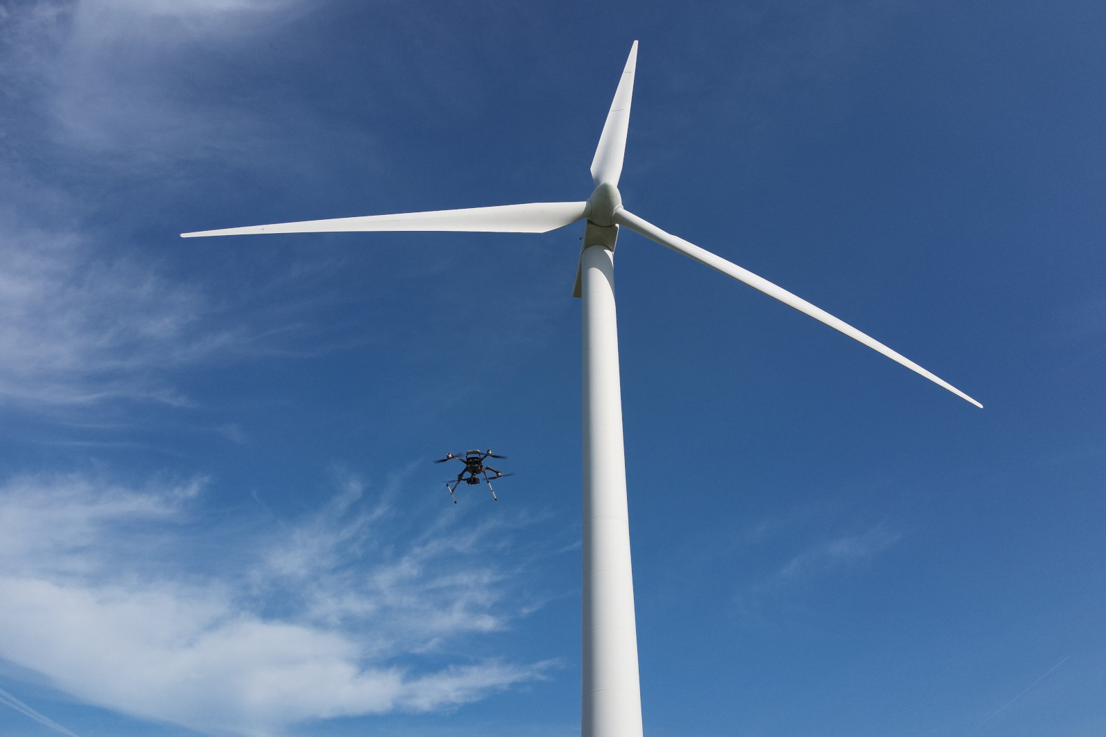 A drone hovering near a large white wind turbine against a clear blue sky with scattered clouds.