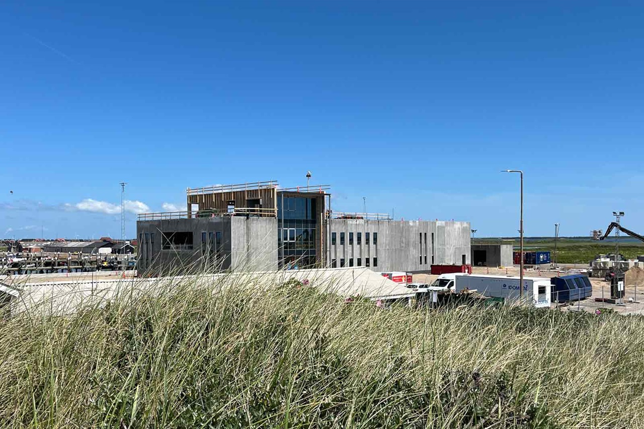 En byggeplads med en moderne bygning i beton og glas under konstruktion ved havet. Græsser i forgrunden.