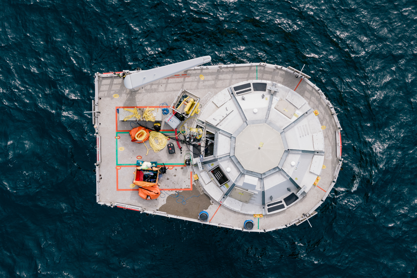 An aerial view of a floating platform equipped with yellow cables, equipment, and safety gear surrounded by deep blue ocean water.