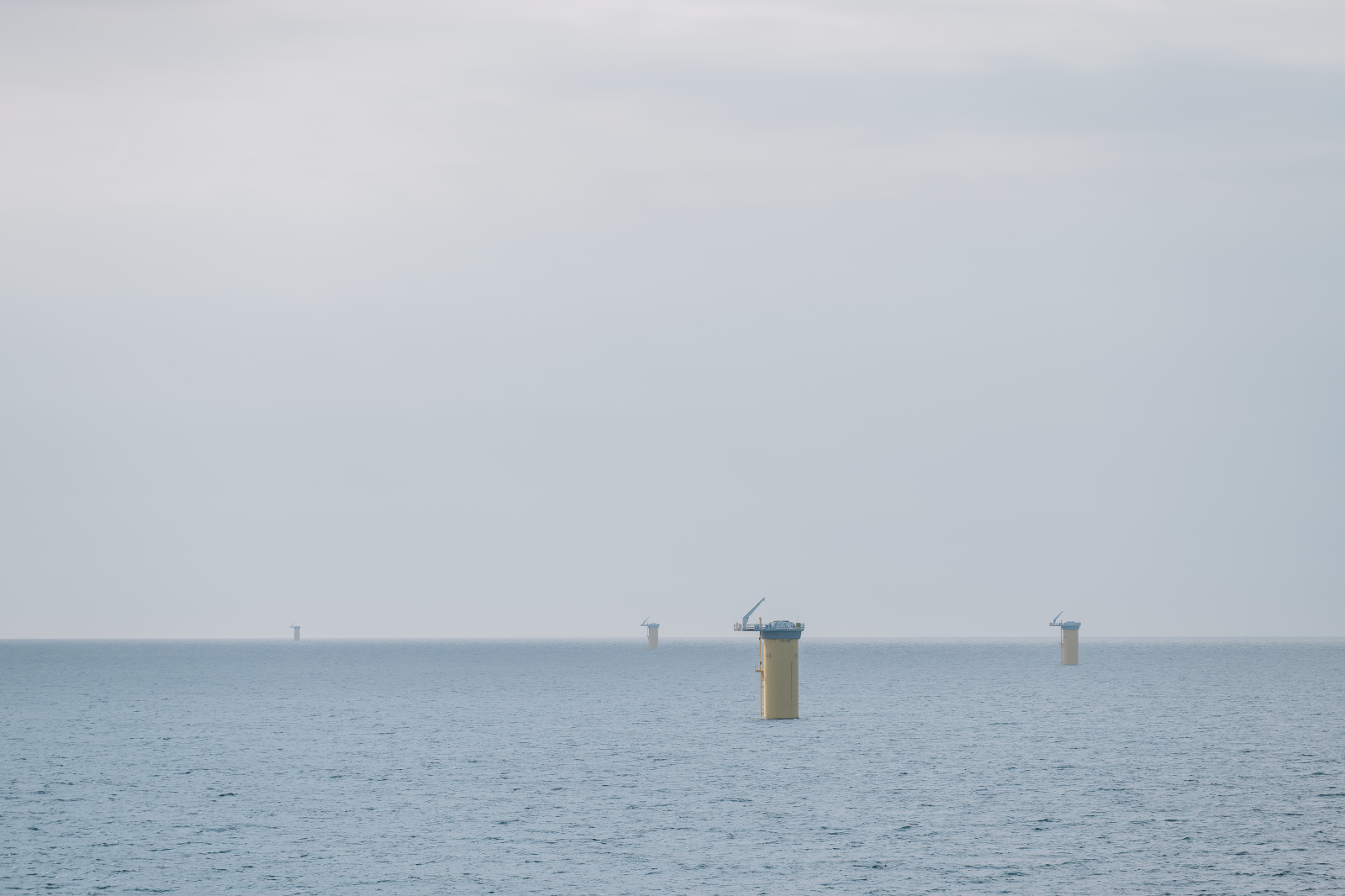 Four yellow offshore wind turbine foundations rise from the calm blue sea under a grey sky.