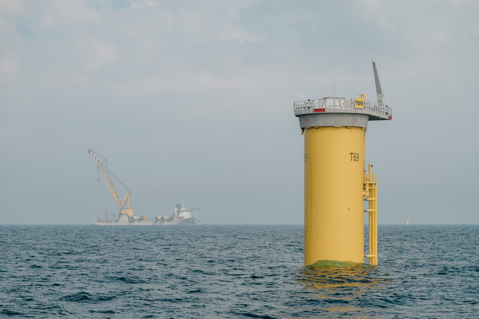 A yellow offshore platform marked T69 stands in calm waters, with a crane ship visible in the background under a cloudy sky.