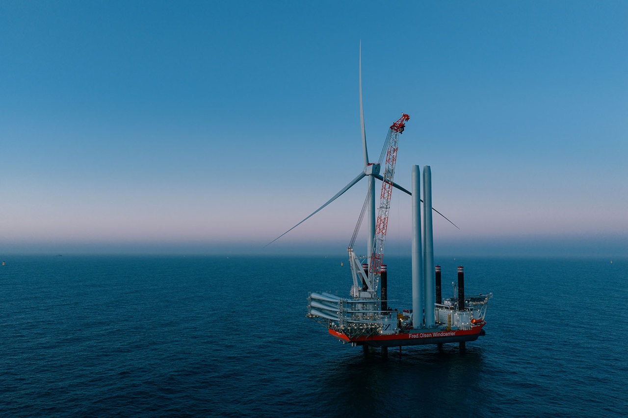 Et offshore installationsskib med en kran løfter en vindmølleklinge ved siden af opførte tårne på stille hav i tusmørke.