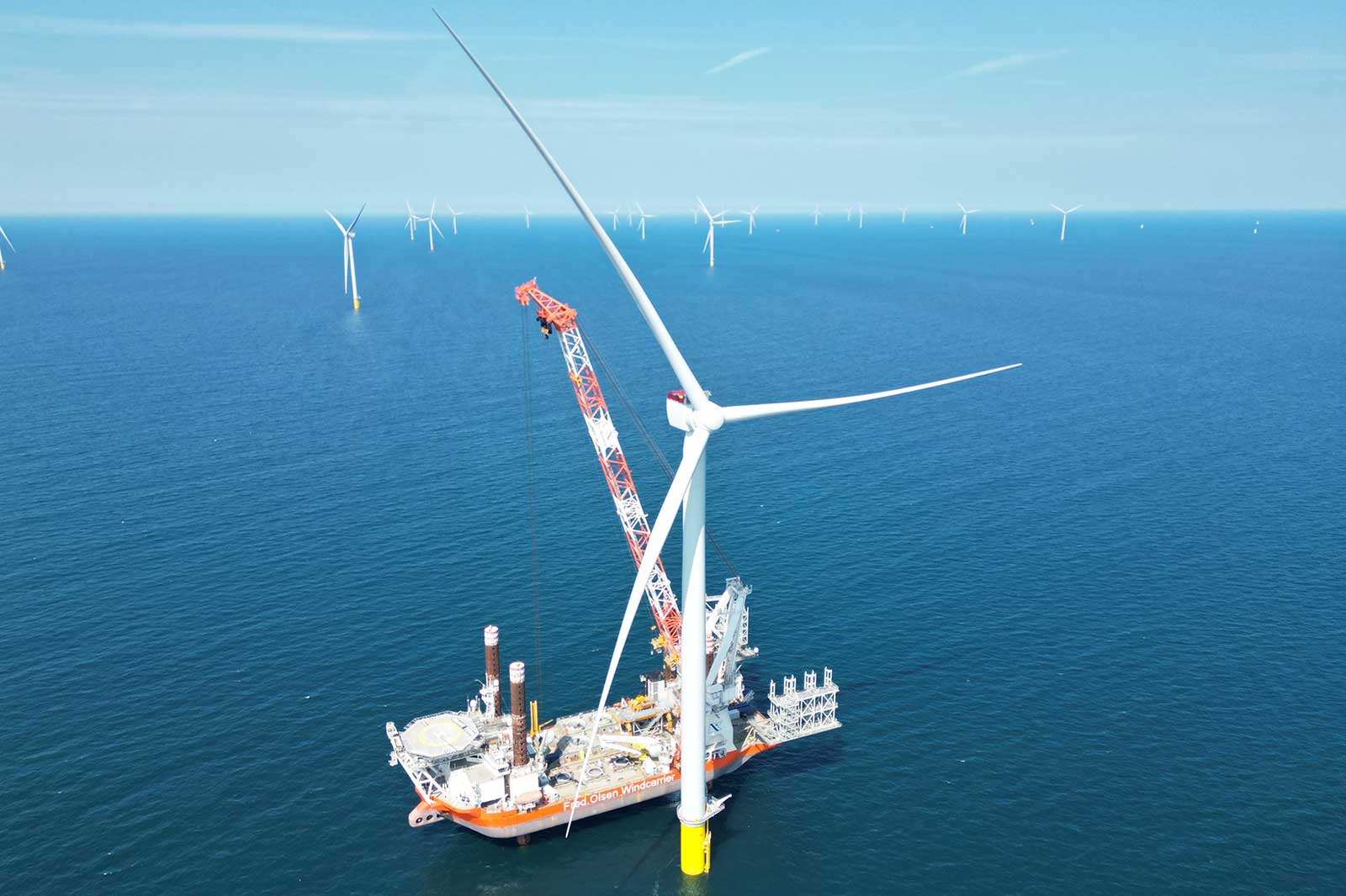 A white offshore wind turbine stands beside an orange installation vessel with a red crane on calm blue sea and clear sky, distant turbines visible.