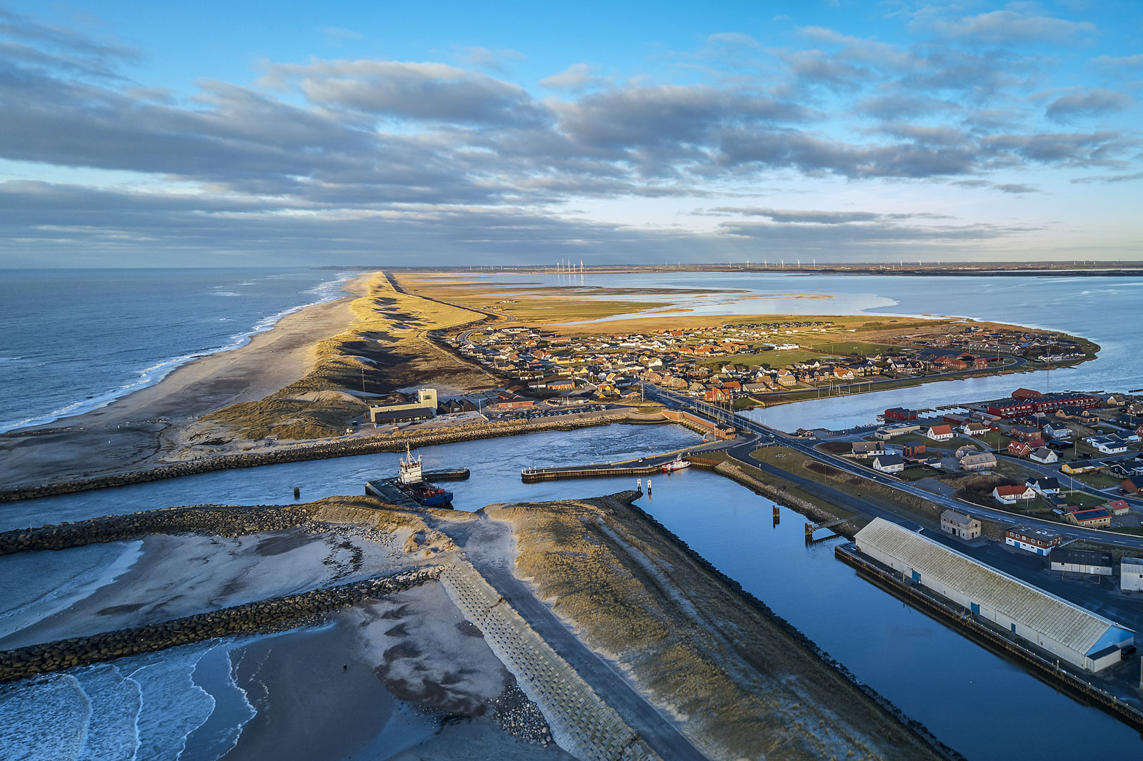 Aerial view of a coastal area featuring a village, a harbour, and a long sandbank, surrounded by water and wind turbines.
