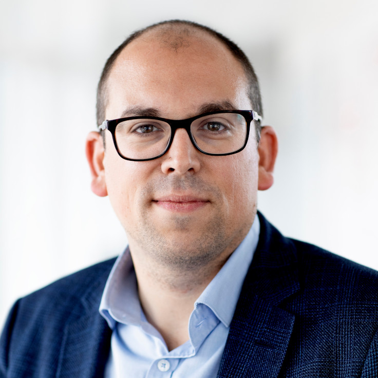 En mand iført en blå skjorte og en mørk jakke står foran en lys baggrund.