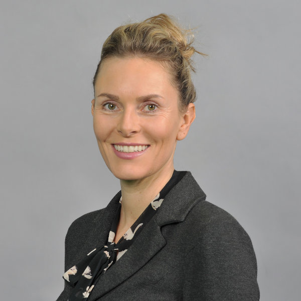 A person in a black blazer with a patterned scarf, standing against a grey background. Portrait of Viola Baumann.